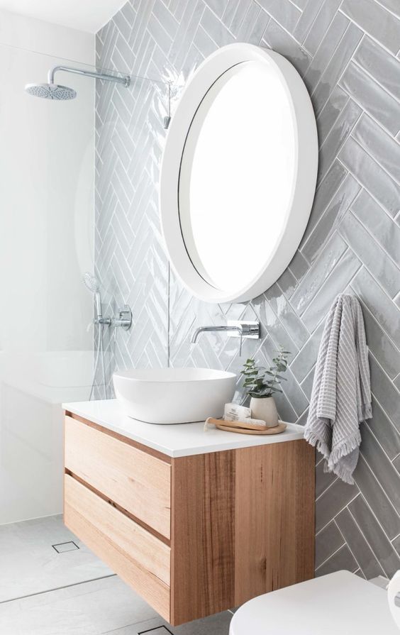 Grey subway tiles arranged in a bold double herringbone pattern behind a round mirror and minimalist timber vanity with white countertop basin.