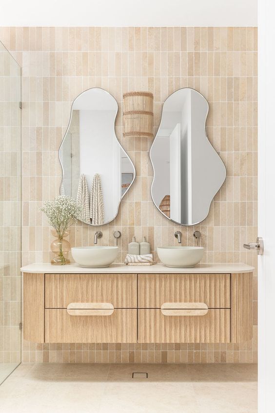 Travertine beige subway tiles laid in a vertical stack pattern behind twin basins and organic-shaped mirrors above a floating timber vanity.