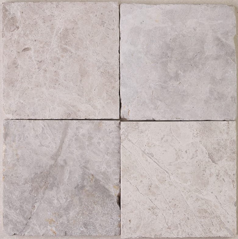 Tumbled square natural stone tiles in mixed neutral tones, laid in a soft grid across an outdoor floor. Visible variation and softened edges.