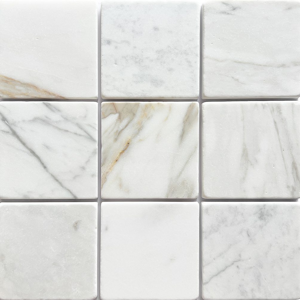 Tumbled square natural stone tiles in mixed neutral tones, laid in a soft grid across an outdoor floor. Visible variation and softened edges.