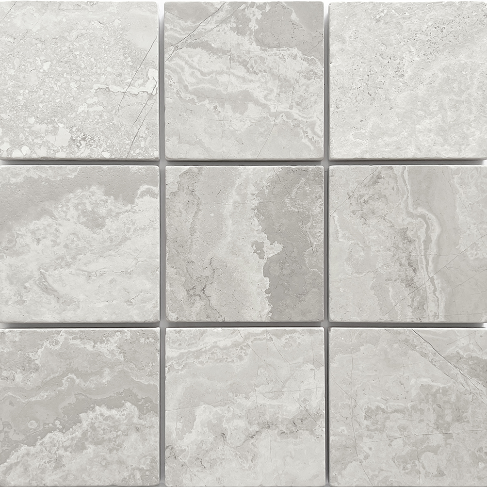 Tumbled square natural stone tiles in mixed neutral tones, laid in a soft grid across an outdoor floor. Visible variation and softened edges.