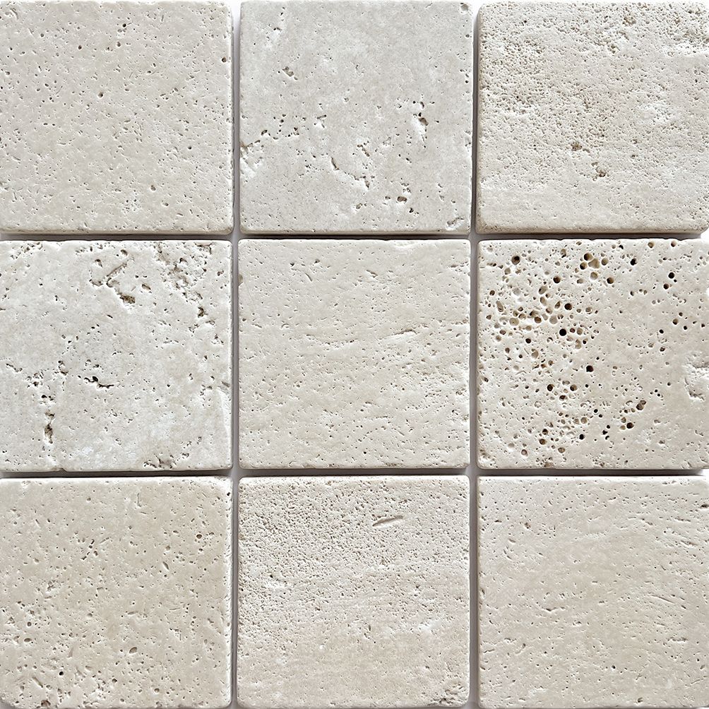Tumbled square natural stone tiles in mixed neutral tones, laid in a soft grid across an outdoor floor. Visible variation and softened edges.
