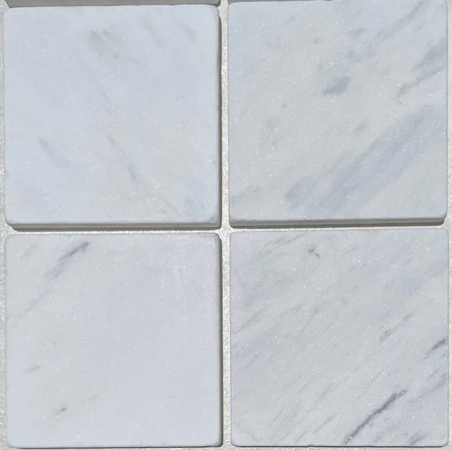 Tumbled square natural stone tiles in mixed neutral tones, laid in a soft grid across an outdoor floor. Visible variation and softened edges.
