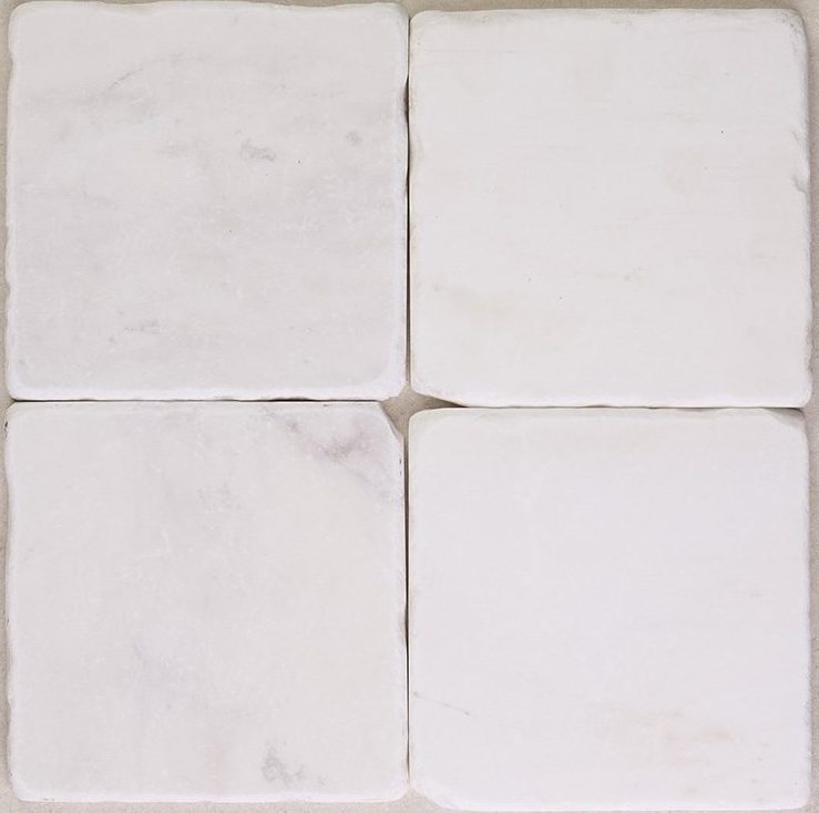 Tumbled square natural stone tiles in mixed neutral tones, laid in a soft grid across an outdoor floor. Visible variation and softened edges.